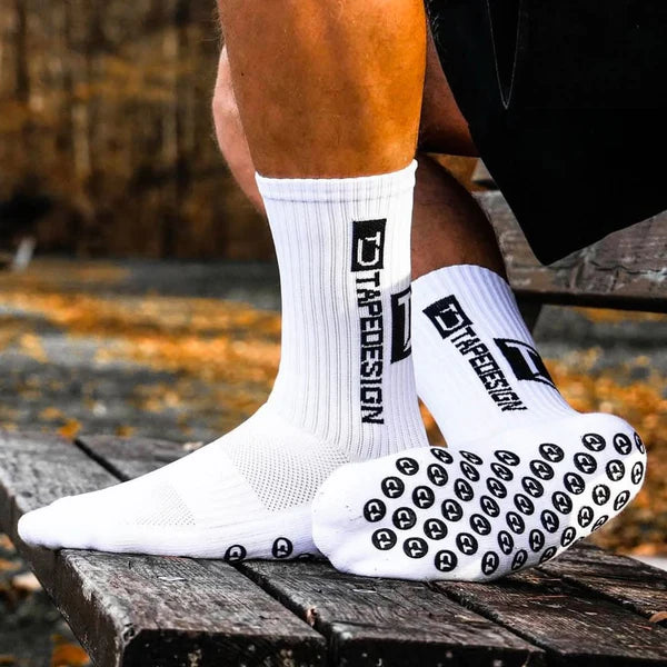 White crew socks with black grip soles and a vertical TapeDESIGN' logo, worn on a person's feet sitting on a wooden bench outdoors.
